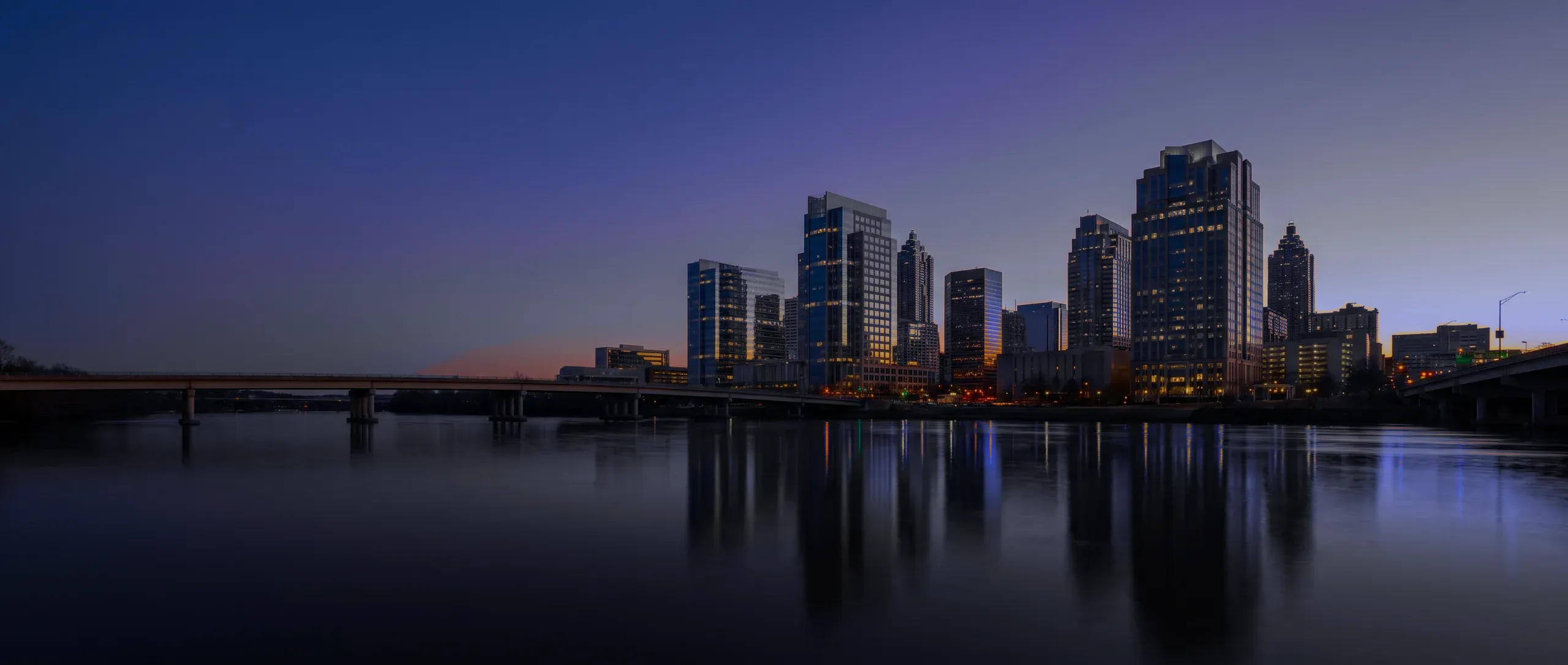 A downtown skyline across a calm river at dusk, a low bridge in the foreground.