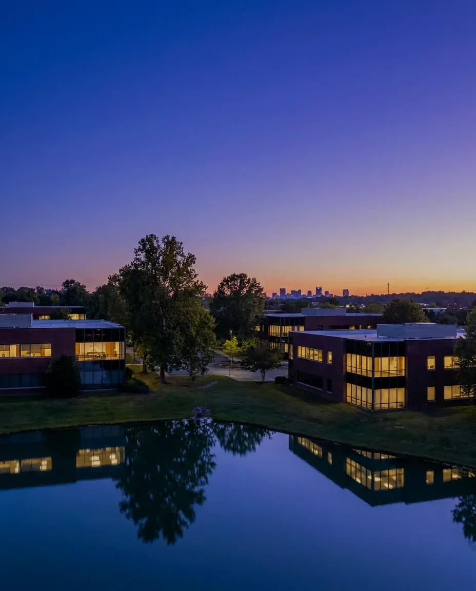 Low office buildings among tall trees at dusk, a city skyline far off.