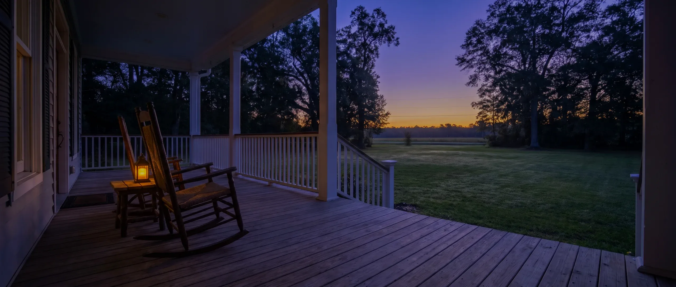 Two empty rocking chairs and a lit lantern on a porch at dusk.